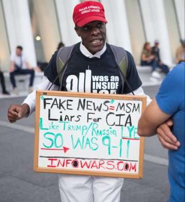 man in black and white shirt with red make America great again cap holding fake news=MSM sign near white building