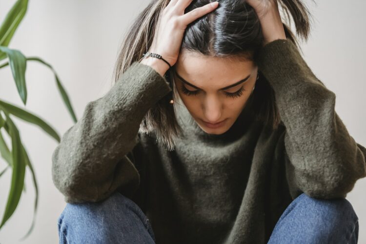 Crop disappointed young ethnic female with dark hair in casual clothes grabbing head with hands and looking down while sitting against white wall at home